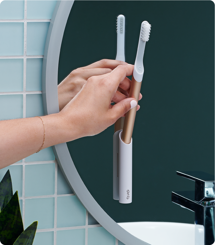 A hand places a white electric toothbrush with a gold handle into a wall-mounted holder on a bathroom mirror, with a second toothbrush already docked beside it against a green and tiled background.