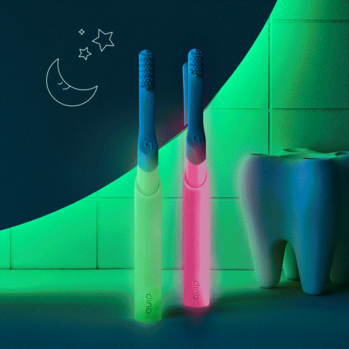 Two quip Kids Sonic toothbrushes in glow-in-the-dark green and pink standing upright on a bathroom counter, with a large tooth model and moon-and-stars illustration in the background.