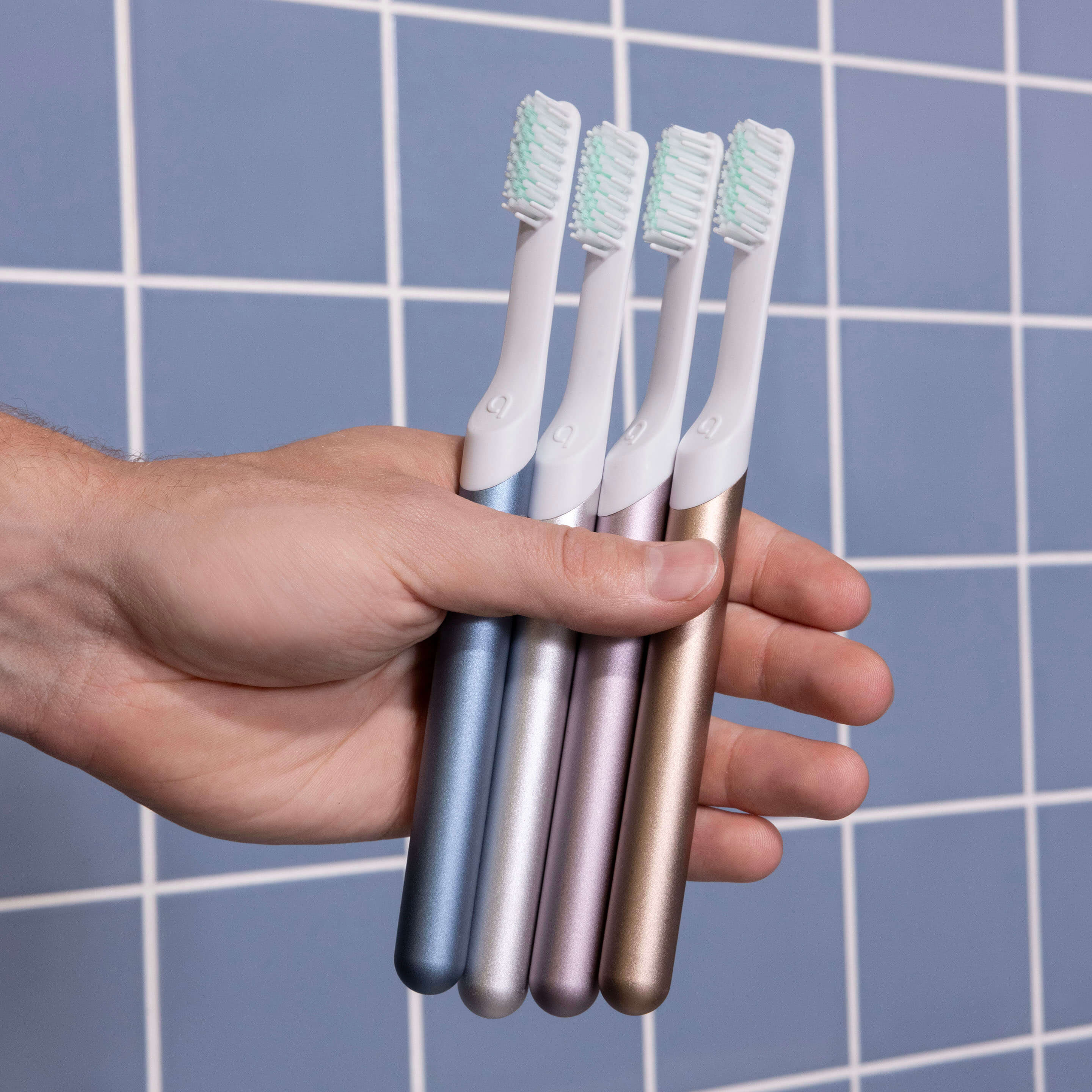 Hand holding four toothbrushes with metallic handles against a tiled bathroom wall.