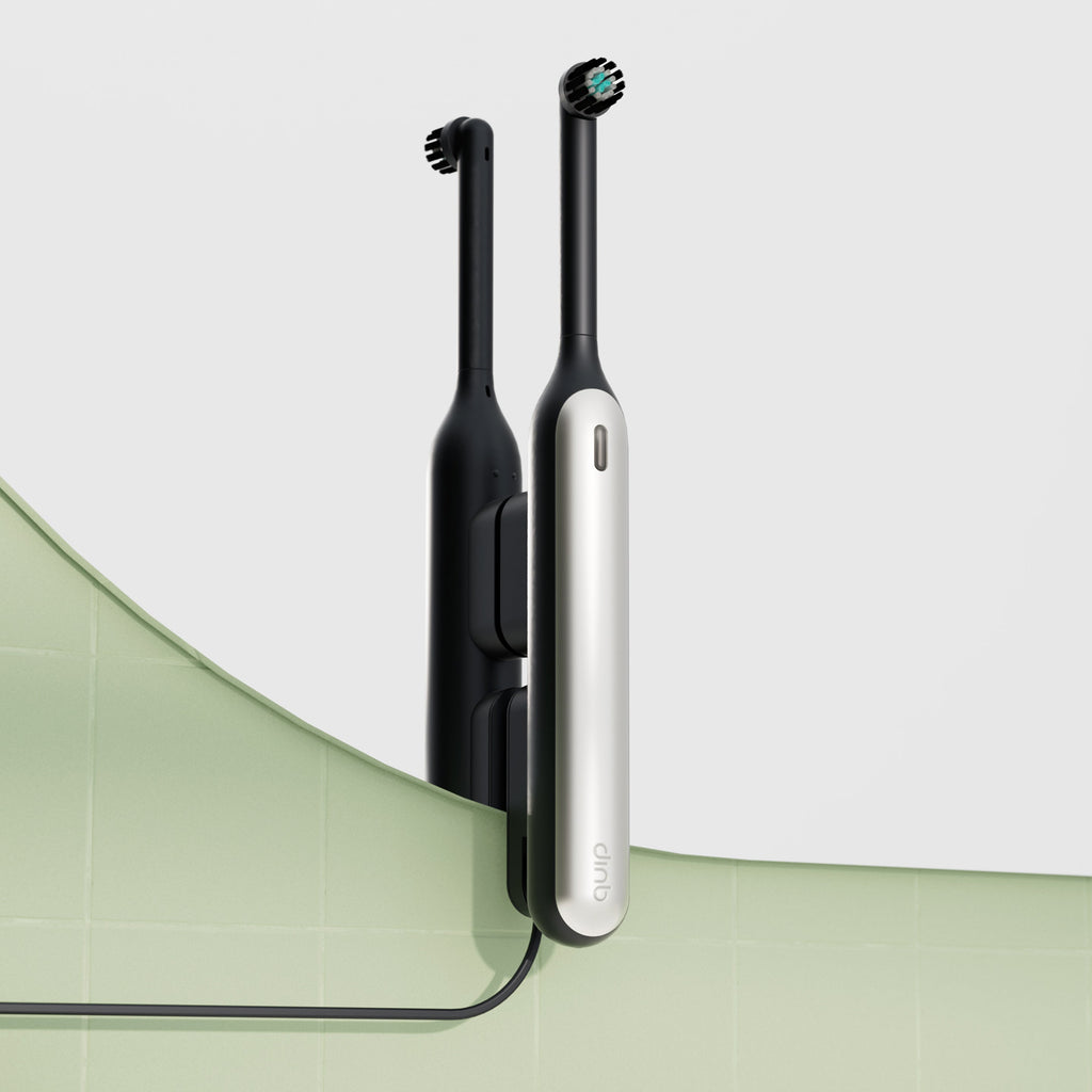Black Rev oscillating toothbrush mounted on a mirror using a black Rev mirror mount, with its charging cable running down across a light green tiled wall.