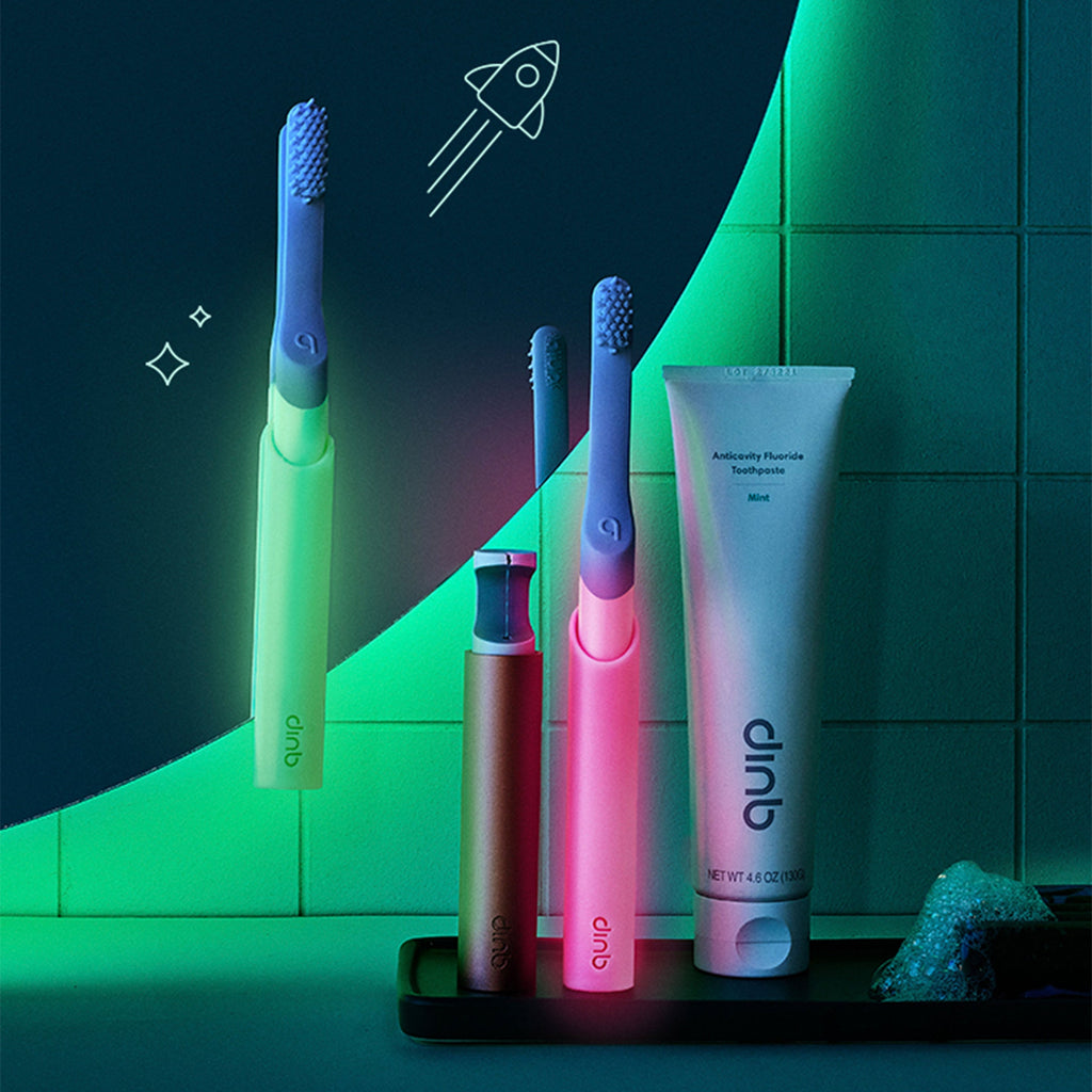 Glow-in-the-dark green and pink quip Kids Sonic toothbrushes displayed with a rinse cup, a tube of anticavity fluoride toothpaste, and playful space-themed illustrations on a bathroom counter.