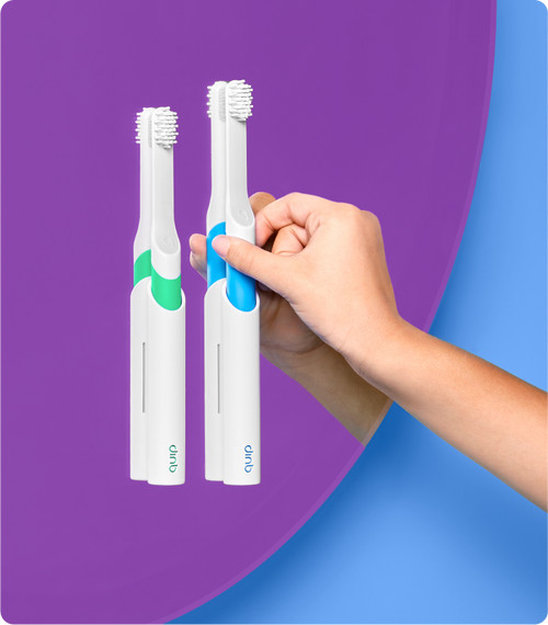 a green and a blue quip kids sonic toothbrush mounted on the bathroom mirror, with a model reaching for the blue brush