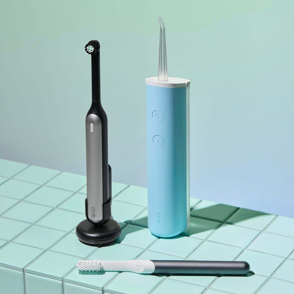 Quip dental tools including a black electric toothbrush, blue water flosser, and white brush on a green tiled surface.