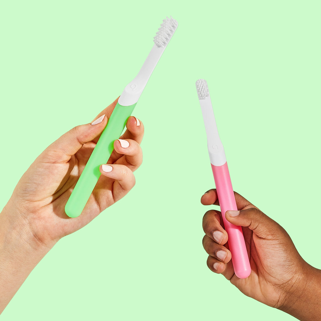 Two hands holding quip Kids Sonic toothbrushes, one green and one pink, against a light green background.