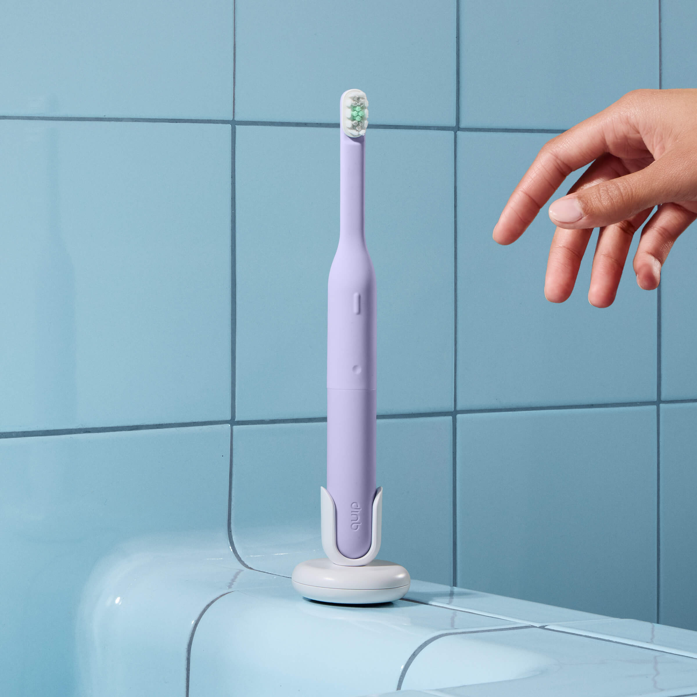 Purple electric toothbrush on a stand with a hand reaching towards it against a blue tiled wall.