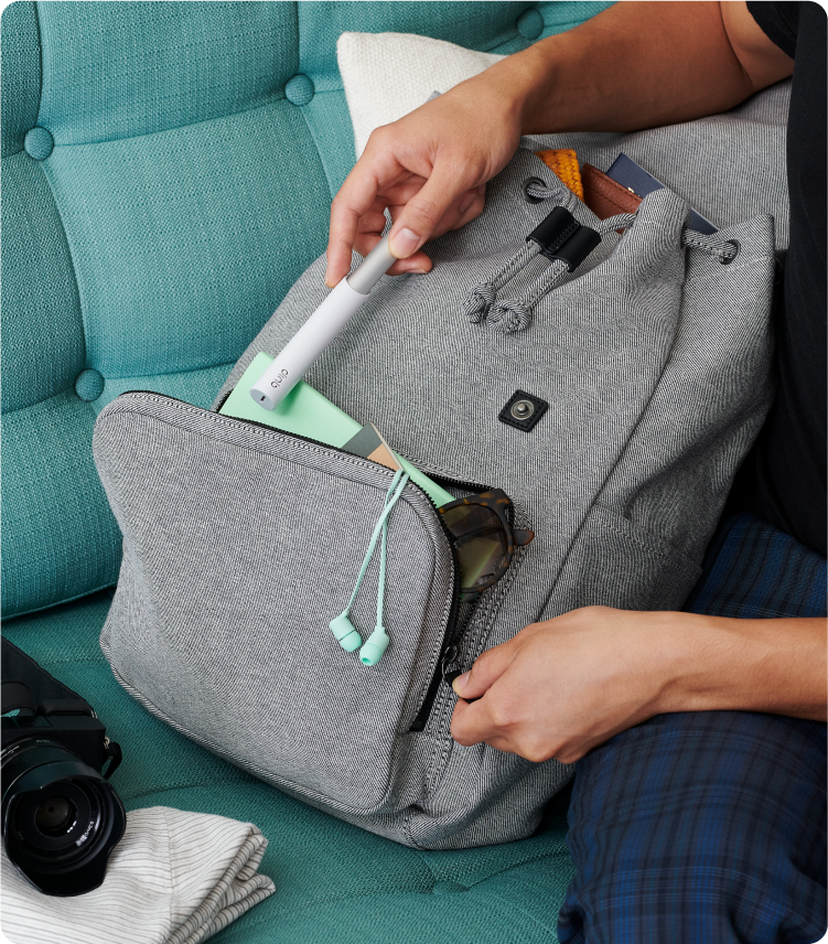 A person places a slim white electric toothbrush into the front pocket of a gray backpack, which contains sunglasses, a notebook, and small accessories, while sitting on a teal sofa.