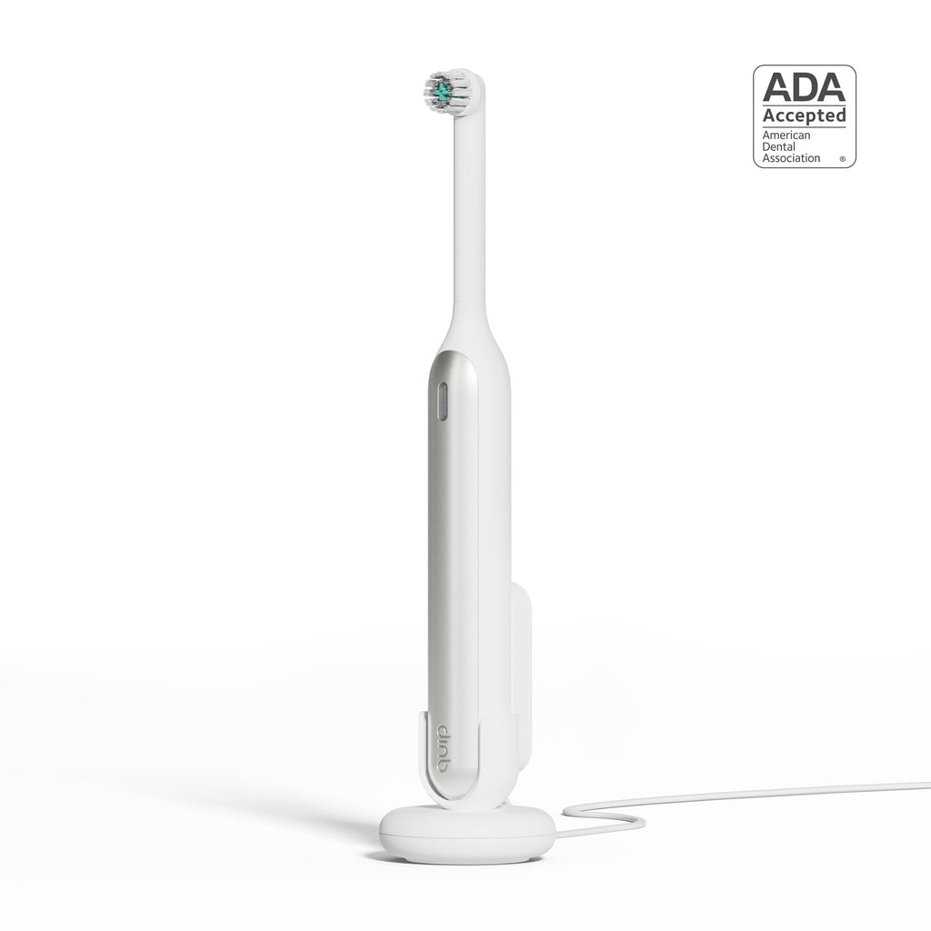 A white quip Rev Electric Toothbrush in the color Silver Cloud on its magnetic charging base, attached to a wall mount, with an ADA Accepted logo in the top right corner.