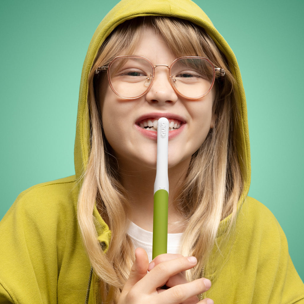 Person wearing a yellow hoodie and glasses, holding a green and white toothbrush against a green background