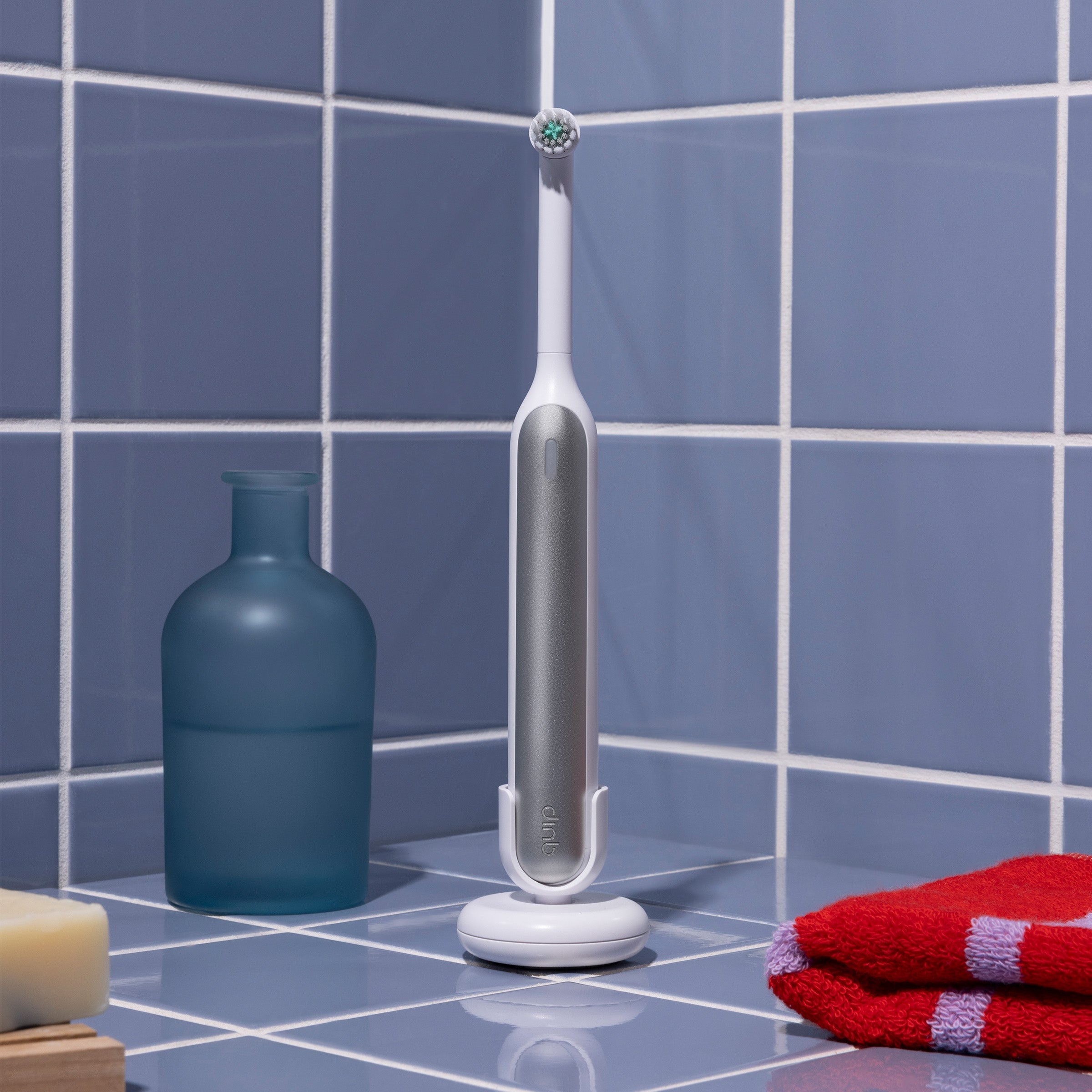 Electric toothbrush on a stand with a blue bottle and red towel on a tiled bathroom floor.