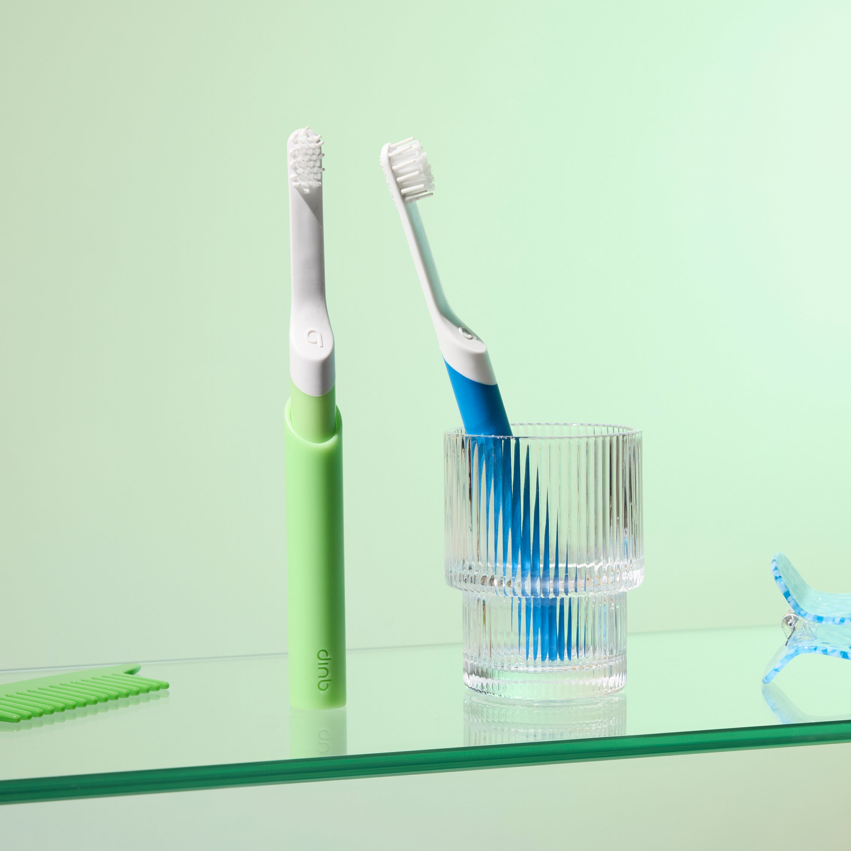 Two toothbrushes displayed on a soft green background—one with a green handle standing upright and another with a blue handle resting inside a clear ridged glass cup, with a green comb placed nearby.